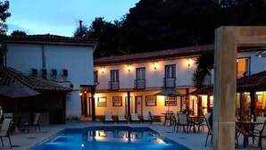 Un espace piscine avec des chaises longues et des parasols, un bâtiment doté de nombreuses fenêtres et d’un espace extérieur aménagé, et un ciel dégagé.