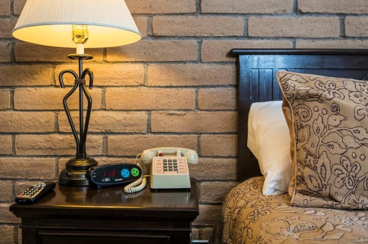 Une chambre d’hôtel avec un mur en briques, une table de chevet en bois, une lampe, un téléphone à cadran, une télécommande et une couvre-lit à motifs.