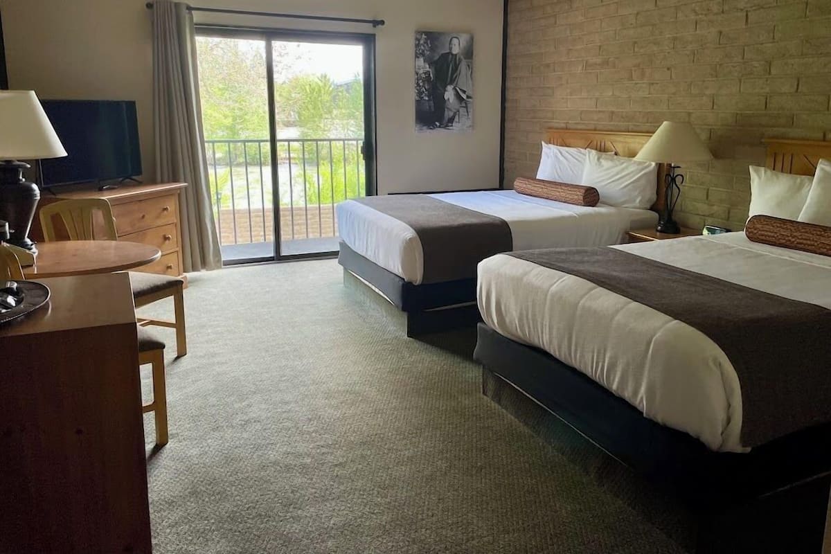 A hotel room with two beds, a desk, a chair, a television, and a balcony with a view of greenery.