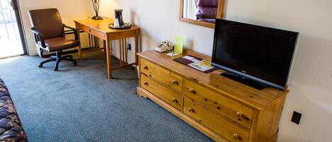 Une chambre d’hôtel avec un lit, un bureau et une chaise, un téléviseur posé sur une commode en bois et une lampe.