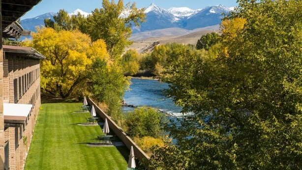 Un hotel a orillas del río con vistas a montañas nevadas, un césped bien cuidado y mobiliario exterior.