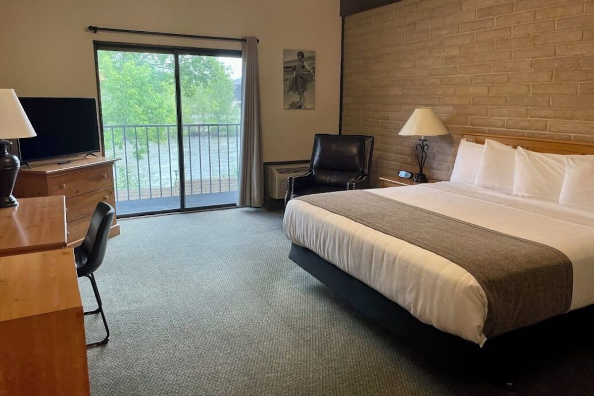 A hotel room with a large bed, a desk with a TV, a chair, a balcony with a view, and a brick wall.