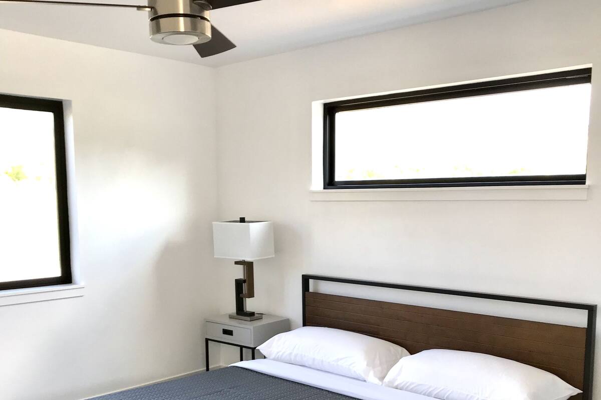 A neatly made bed with a grey textured bedspread, a wooden headboard, and white pillows. There is a nightstand with a lamp, a ceiling fan, and a window with blinds.