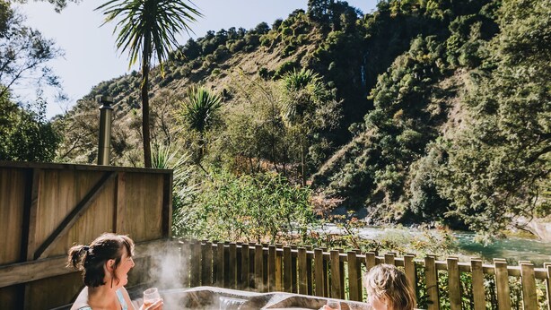 Two people in a hot tub outdoors surrounded by trees and hills. - River Valley Lodge (Pukeokahu)