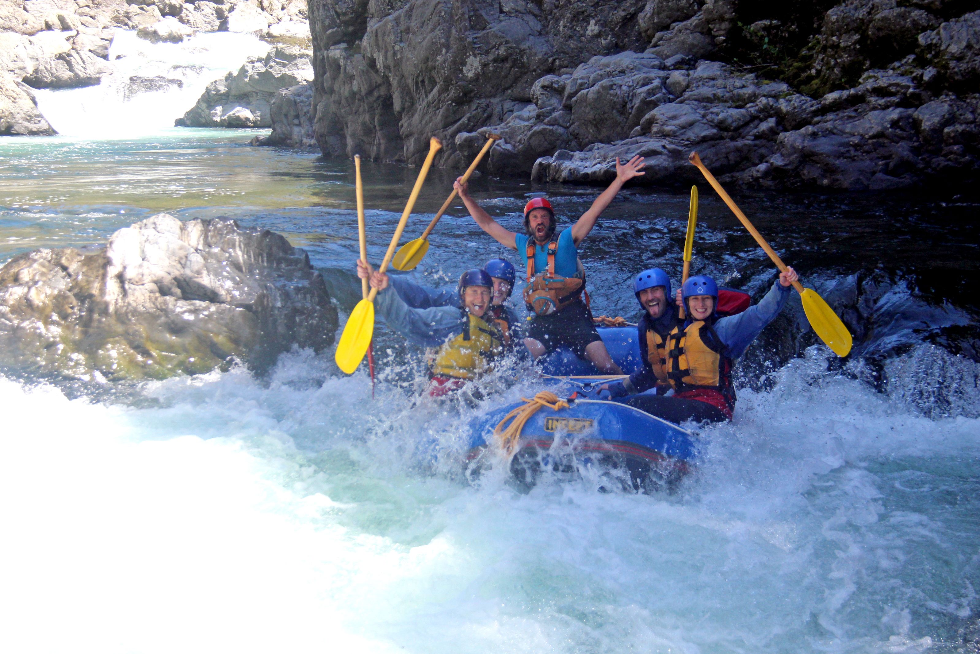 A group of people in a blue raft navigating through a river with rapids, surrounded by rocky cliffs.