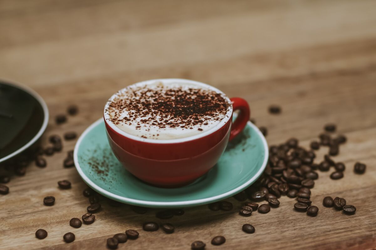 A cup of coffee with a frothy top and a sprinkle of cocoa powder on a red saucer.