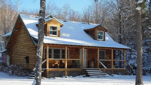 Una baita di legno con veranda e un piccolo balcone superiore, circondata da neve e alberi.