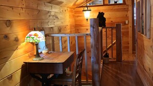 Interno di una baita in legno con un tavolo da pranzo, sedie, una lampada e una scala che conduce a un piano superiore.