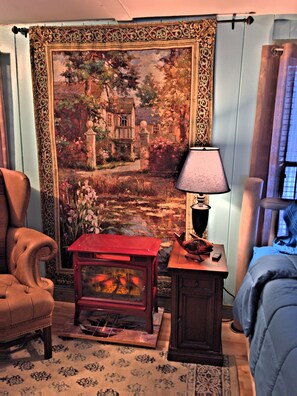 A room with a large tapestry, a red stove, a wooden nightstand, a lamp, a leather armchair, and a bed with a blue blanket. - Causeway Bike Path steps away with Water access and Kayaks. (Colchester)