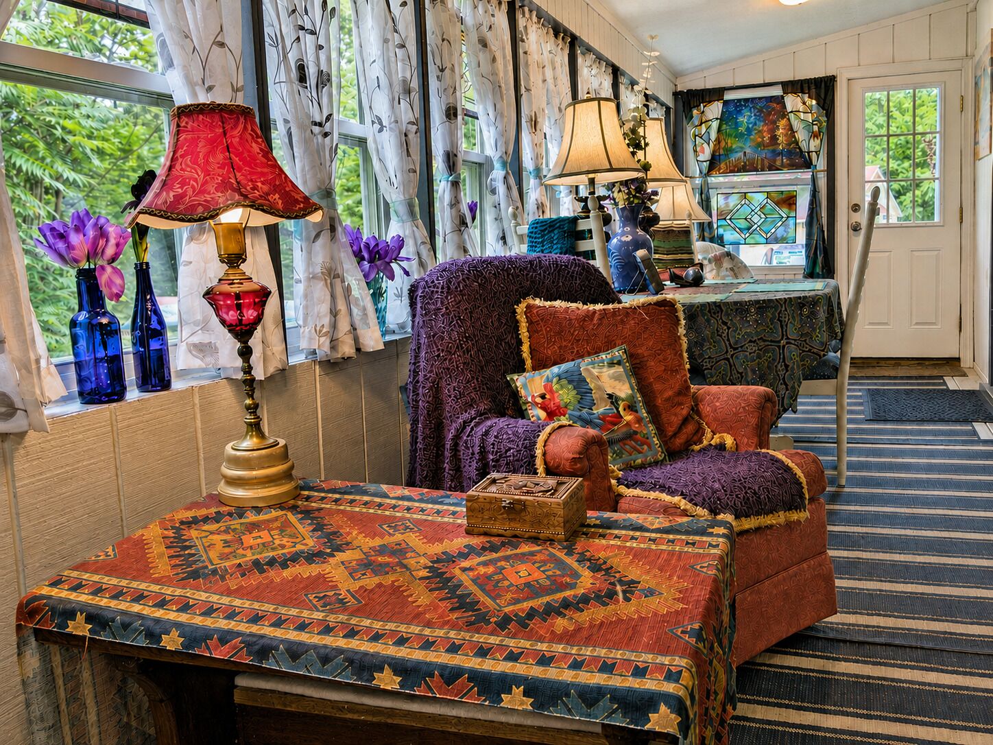 A cozy room with a patterned rug, a chair with a cushion, a table with a lamp, and a window with curtains.