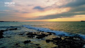 Un paysage côtier avec un coucher de soleil sur l’océan, une côte rocheuse et une masse terrestre visible au loin.
