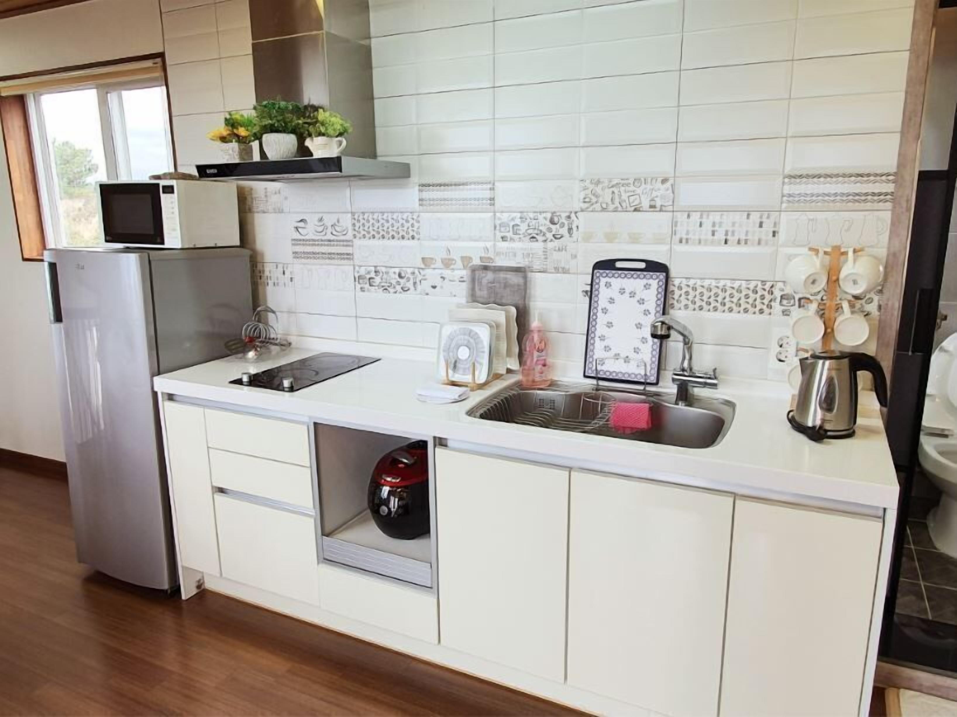 Cozinha moderna com armários brancos, geladeira de aço inoxidável, micro-ondas, pia com chaleira e um ventilador.