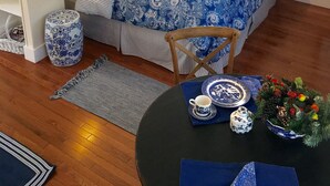 A bedroom with a bed, bedside lamps, a window with blinds, a wooden chair, a round table with a blue tablecloth, and a decorative plant.
