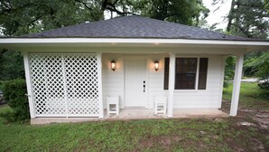 A small, single-story house with a white exterior, a dark roof, and a front porch with two steps. - Detached Cottage In A Rural Woodsy Neighborhood. (Shreveport)