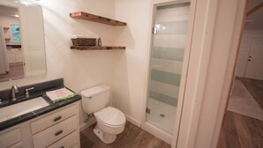 A bathroom with a white toilet, a black sink, and a mirror. There are wooden shelves above the toilet and a glass shower door. - Detached Cottage In A Rural Woodsy Neighborhood. (Shreveport)