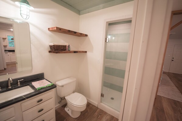 A bathroom with a white toilet, a black sink, and a mirror. There are wooden shelves above the toilet and a glass shower door.