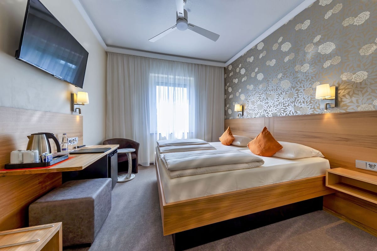 Habitación de hotel con cama, mesitas de noche, escritorio con cafetera, una silla, televisor y ventilador de techo.