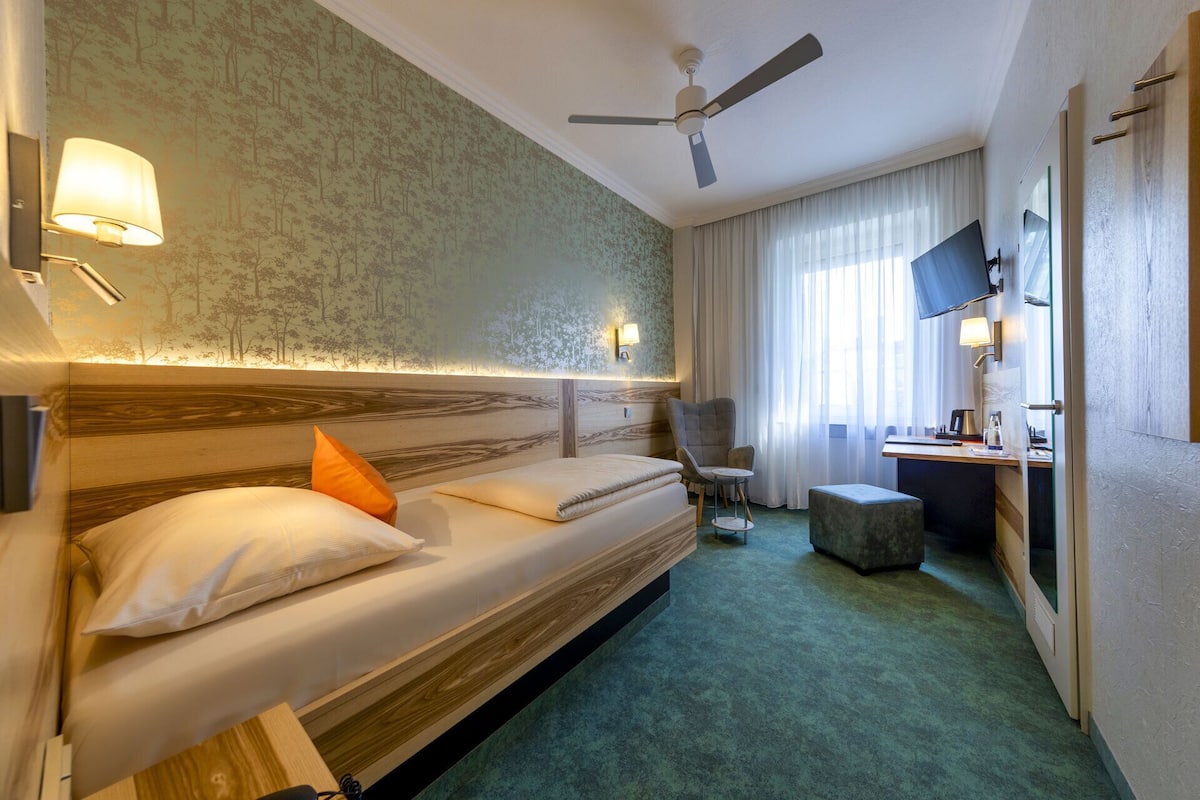 Una habitación de hotel con cama, escritorio, silla, mesita, ventilador de techo y ventana con cortinas.