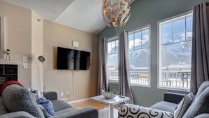 A modern living room with grey sofas, a white coffee table, and a flat-screen TV mounted on the wall. Large windows offer a view of mountains and a balcony. - Summit Place 2B | Excellent Location, Amazing Views (Canmore)