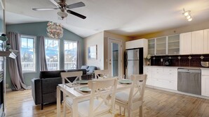 A modern kitchen with white cabinets, a refrigerator, and a dining area with a table and chairs. - Summit Place 2B | Excellent Location, Amazing Views (Canmore)