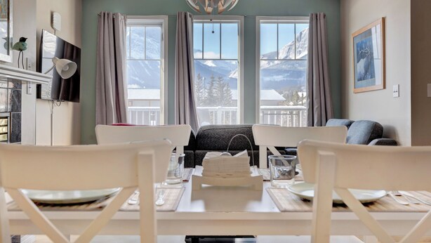 A dining area with a table set for two, a chandelier, and a view of mountains through large windows. - Summit Place 2B | Excellent Location, Amazing Views (Canmore)