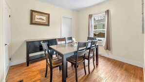A dining room with a wooden table and chairs, a bench, a framed picture on the wall, and a window with curtains. - Left Field - All Star Baseball Rentals (Oneonta)
