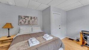 A neatly made bed with a beige comforter and two folded towels, a painting of flowers on the wall, a lamp on a bedside table, and a door to the right. - Left Field - All Star Baseball Rentals (Oneonta)