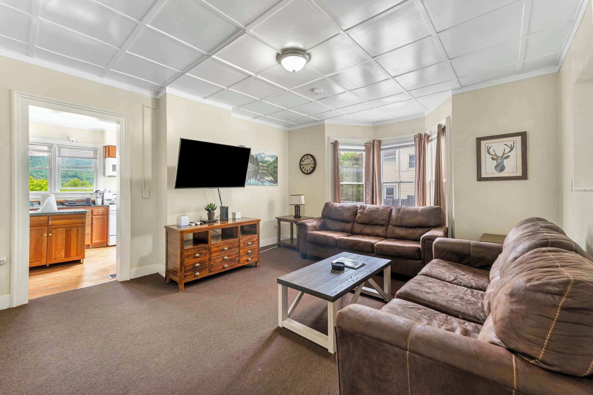 A spacious living room with a brown leather sofa, a coffee table, and a flat-screen TV mounted on the wall. There is a wooden cabinet and a small table with a lamp.