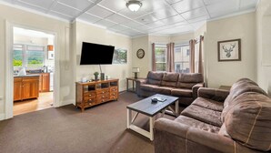A spacious living room with a brown leather sofa, a coffee table, and a flat-screen TV mounted on the wall. There is a wooden cabinet and a small table with a lamp. - Left Field - All Star Baseball Rentals (Oneonta)