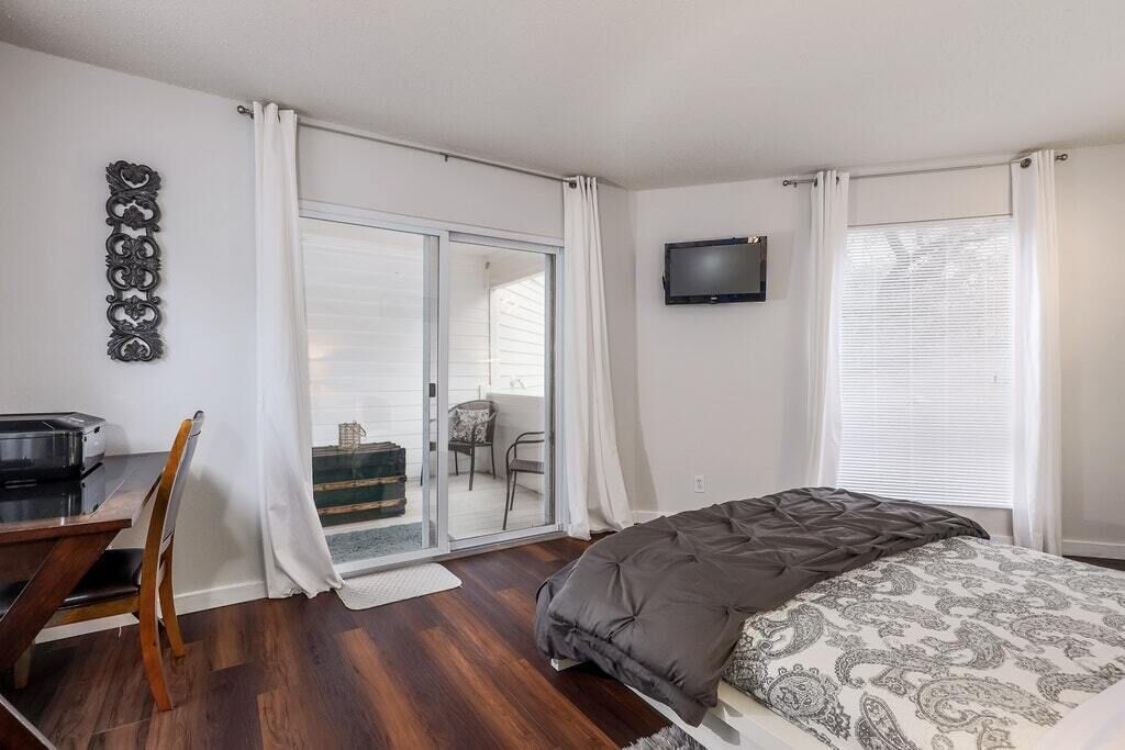 A bedroom with a bed, a desk, a chair, a TV, and a sliding glass door leading to a balcony.
