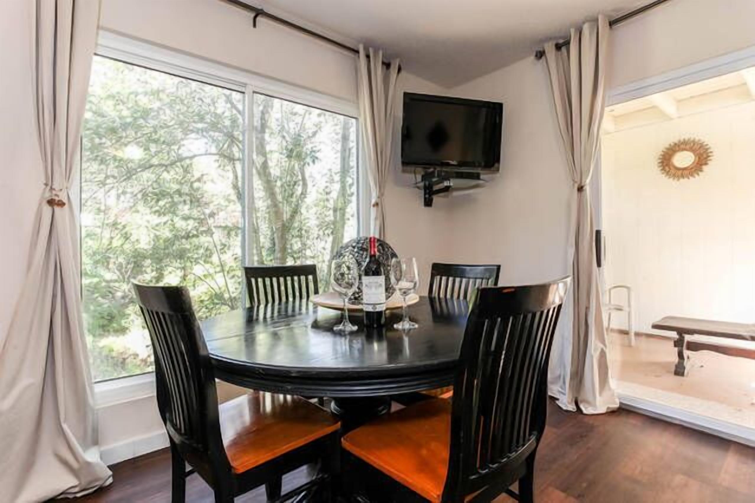 Zona de dinar amb taula rodona, cadires, televisió penjada a la paret i vista a arbres a través de grans finestres.