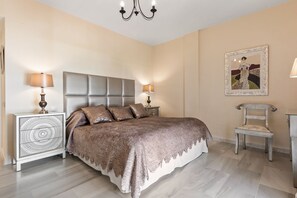 A bedroom with a large bed, two bedside tables, a chair, a chandelier, and a framed picture on the wall. - Beachside Puerto Banus Stunning Apartment 212 (Marbella)