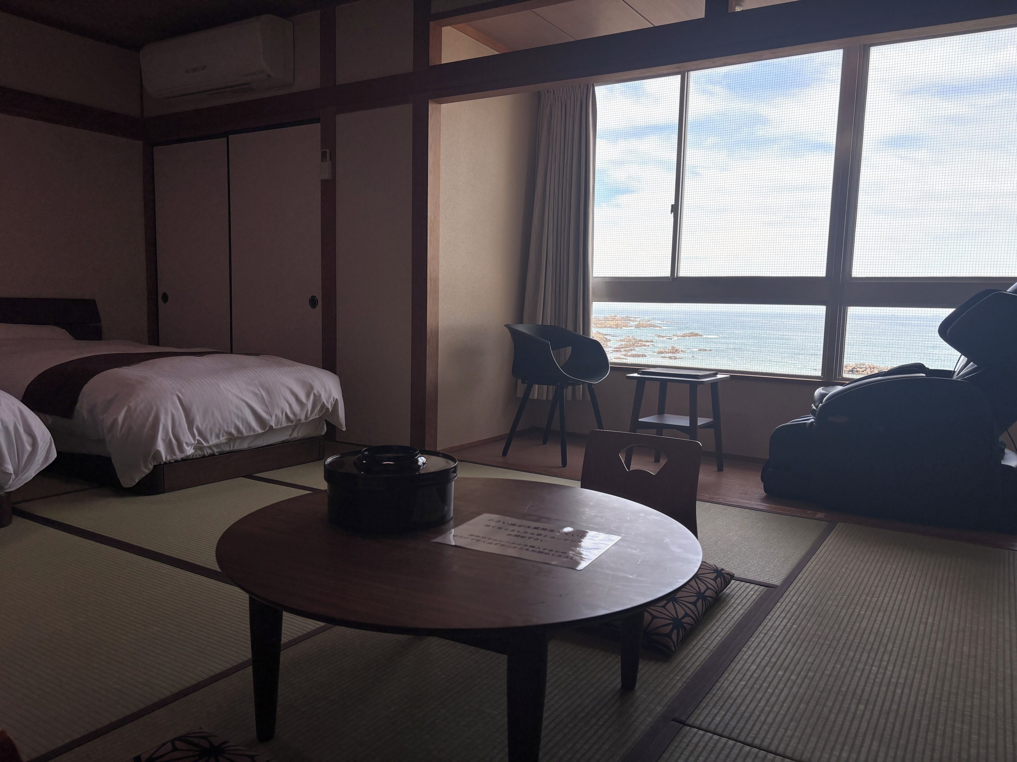 A hotel room with a bed, a table with a menu, a chair, and a view of the sea through large windows.