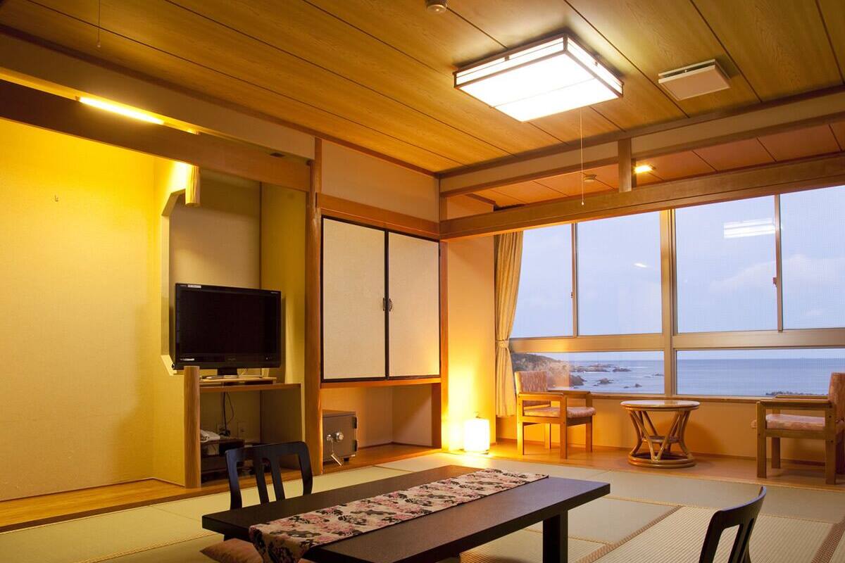 Una habitación con techo de madera, televisor, mesa con posavasos y vista al mar a través de amplias ventanas.