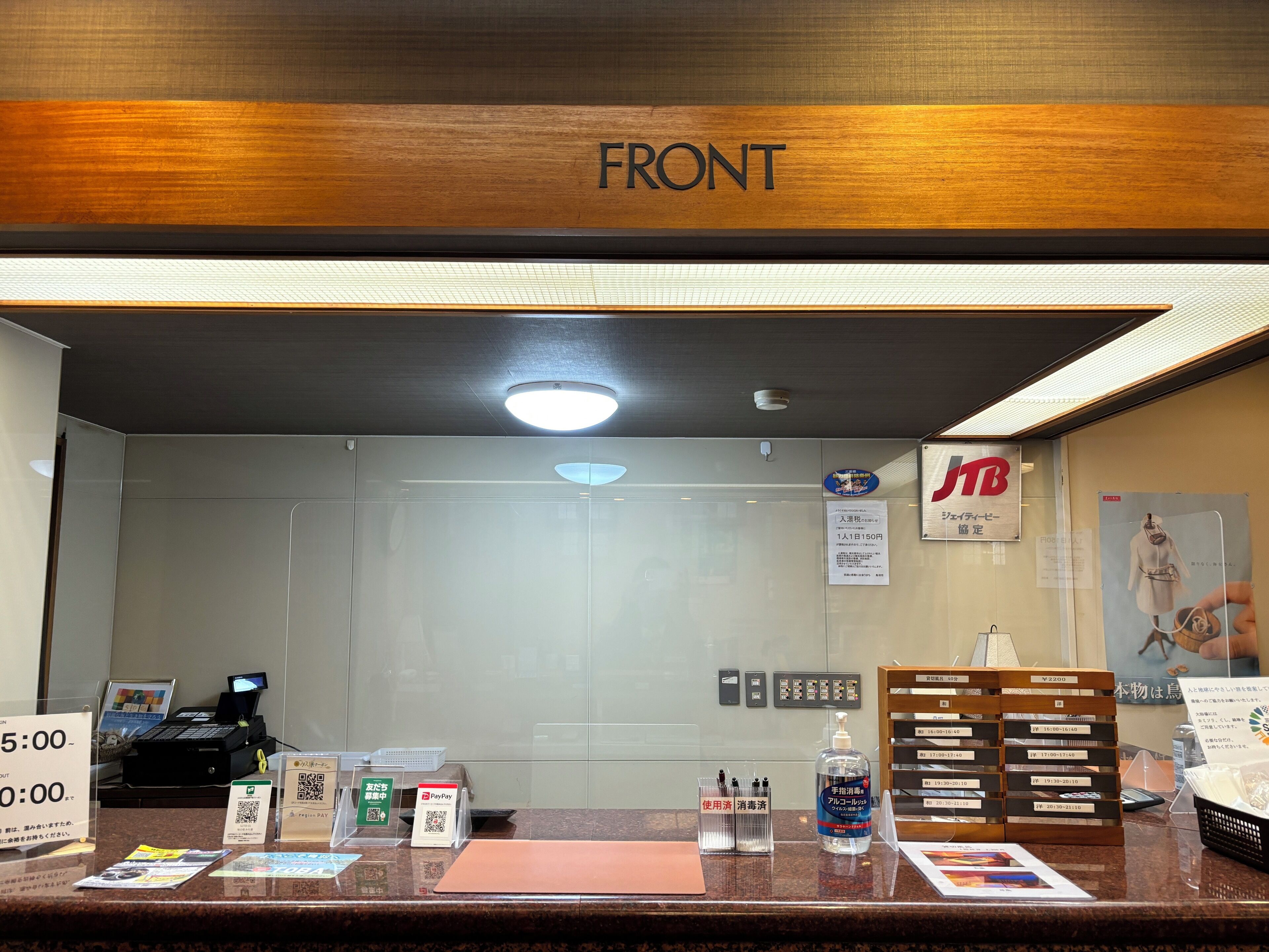 Una mesa de recepción con un letrero que dice 'FRONT', una pantalla digital, un letrero 'TB', un código QR y un lector de tarjetas PayPay.