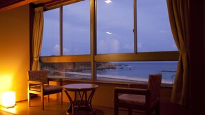 Quarto com grande janela com vista para uma praia, duas cadeiras, uma mesa pequena e um abajur.