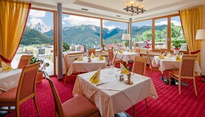 A restaurant with tables set for dining, large windows offering mountain views, and a red carpeted floor.