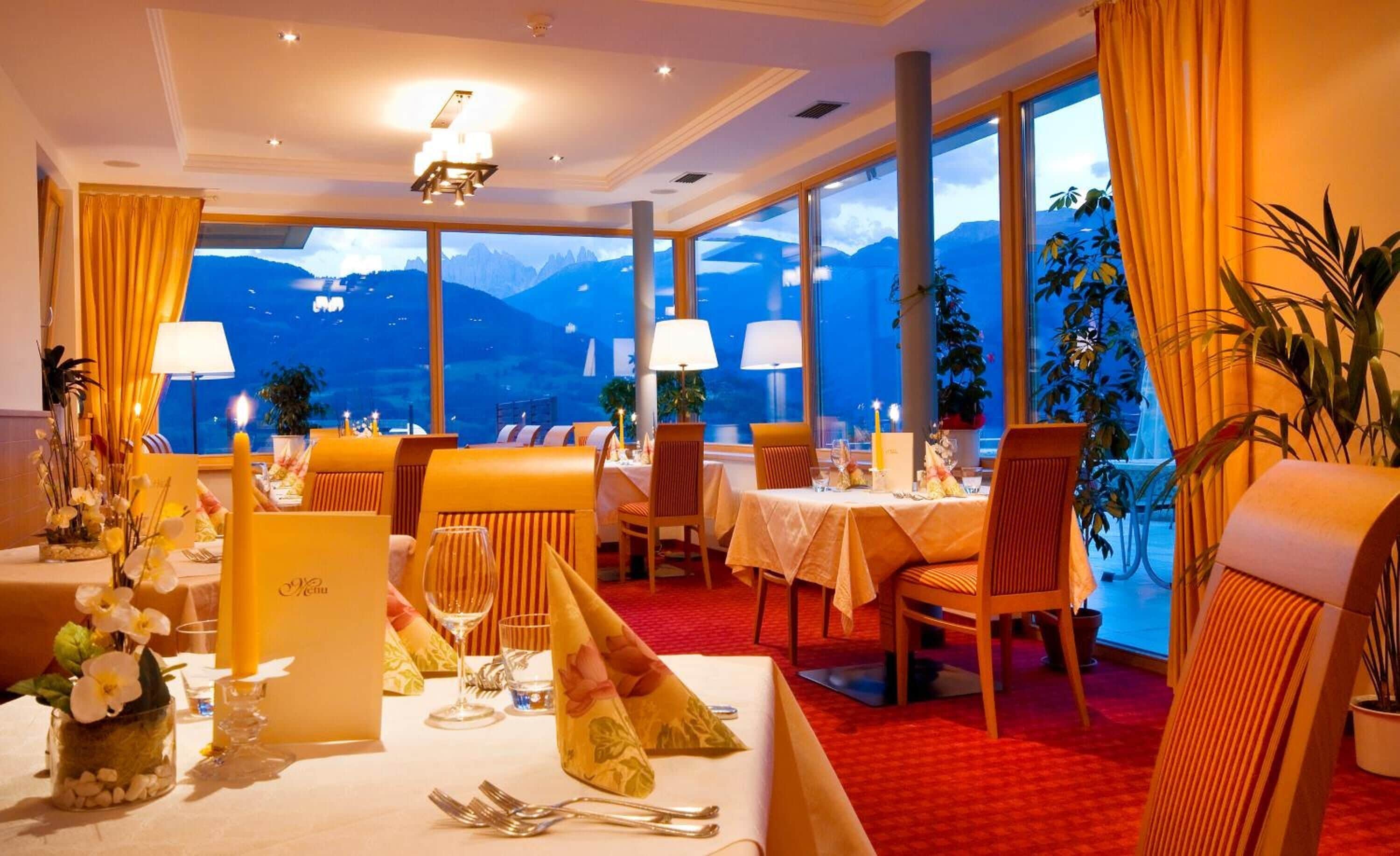 Un ristorante con tavoli apparecchiati per la cena, ampie finestre con vista sulle montagne e illuminazione calda.