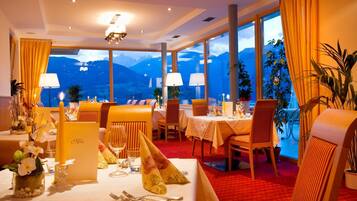 Un ristorante con tavoli apparecchiati per la cena, ampie finestre con vista sulle montagne e illuminazione calda.
