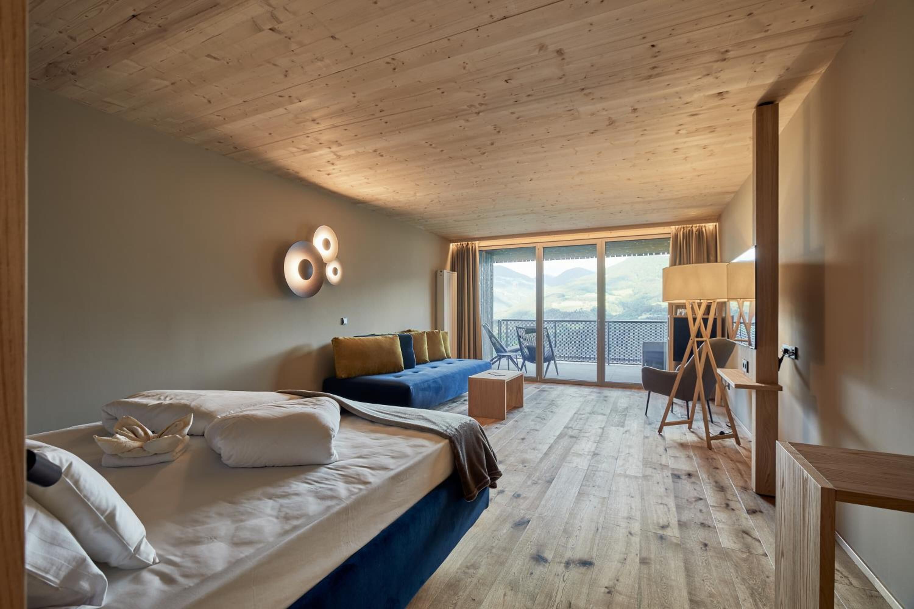 Ein Hotelzimmer mit Holzbalkendecke, einem Bett mit weißer und brauner Bettwäsche, einer blauen Couch, einem kleinen Tisch, einem Stuhl und einem großen Fenster mit Blick ins Freie.