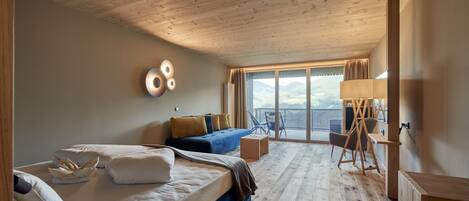 A hotel room with a wooden ceiling, a bed with white and brown bedding, a blue sofa, a small table, a chair, and a large window with a view of the outdoors.