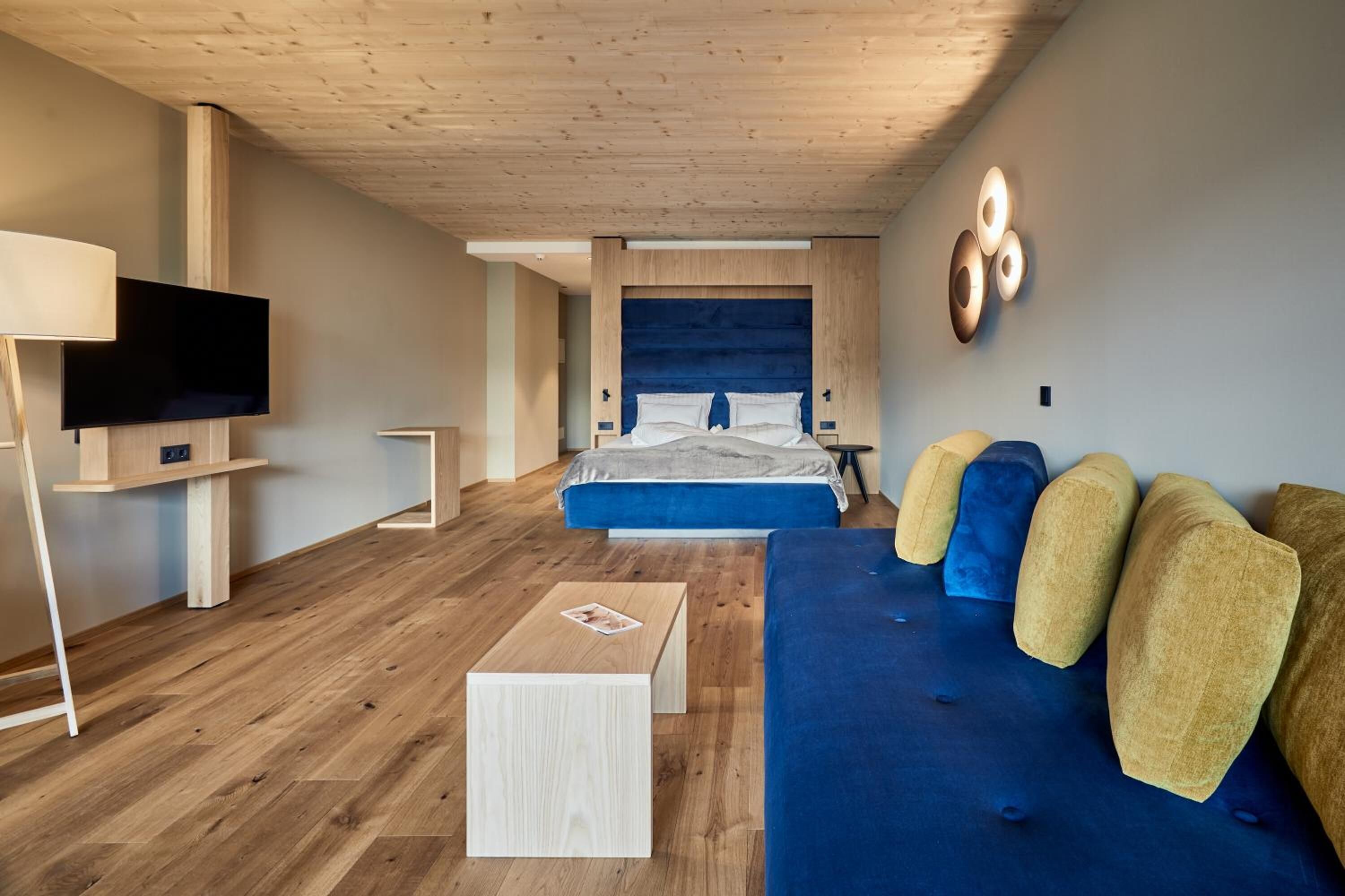 Ein modernes Hotelzimmer mit Holzboden, einem großen Bett mit blauer Bettwäsche, einem blauen Sofa mit Kissen sowie einem an der Wand befestigten Flachbildfernseher und einem kleinen Nachttisch.
