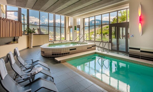 A modern indoor pool area with a hot tub, seating, and large windows offering mountain views.
