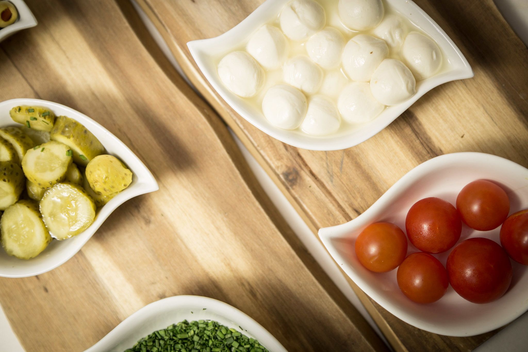 A wooden table with three white dishes containing pickles, boiled eggs, and tomatoes.