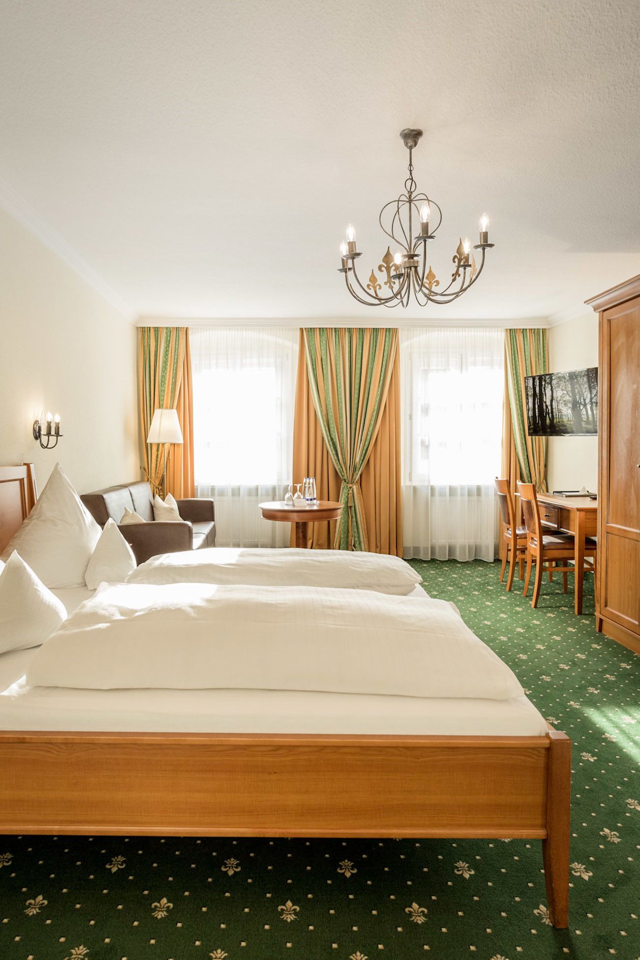 A hotel room with a large bed, a desk with chairs, a television, and a chandelier.