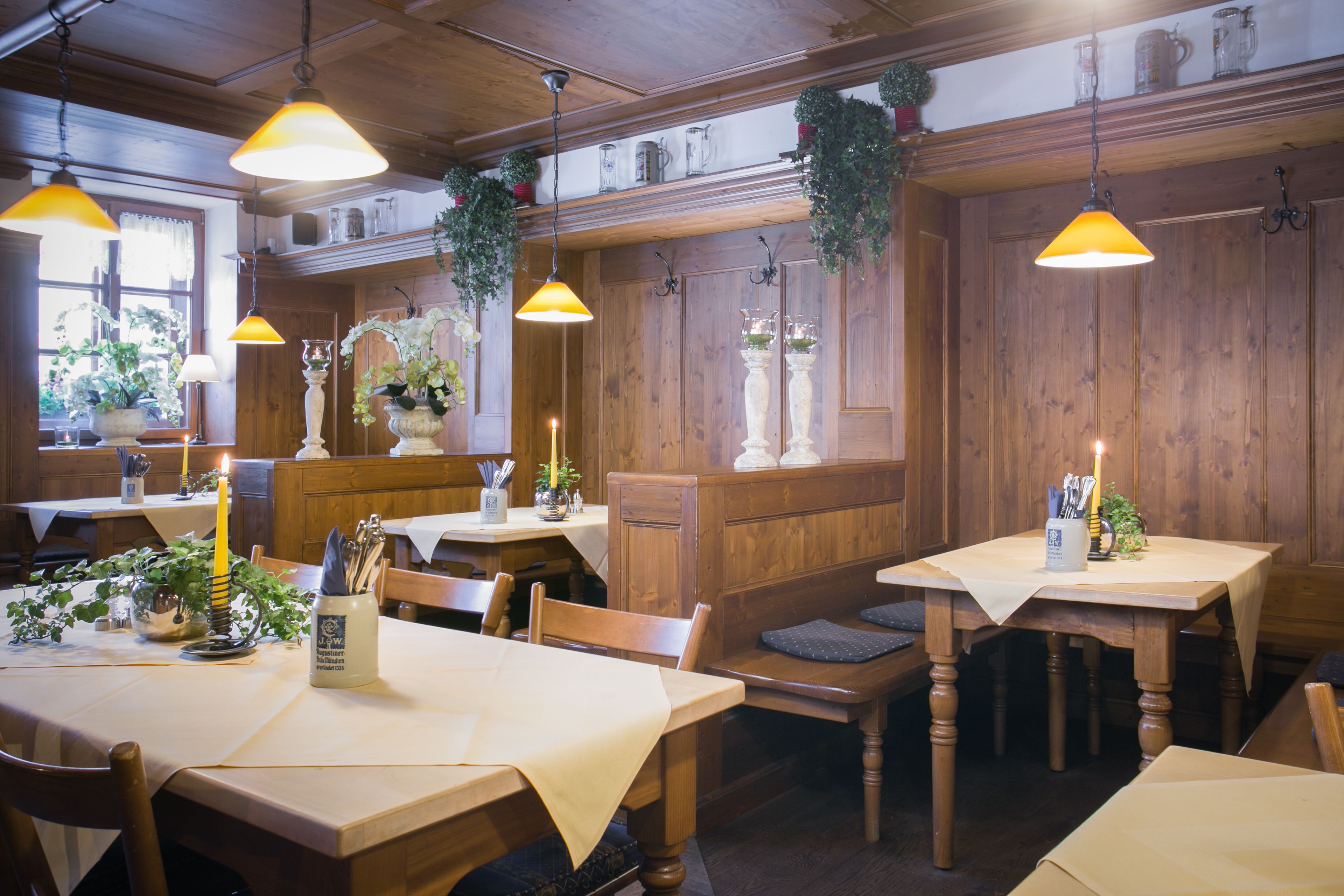 A restaurant interior with wooden paneling, tables with white tablecloths, hanging plants, and pendant lighting.