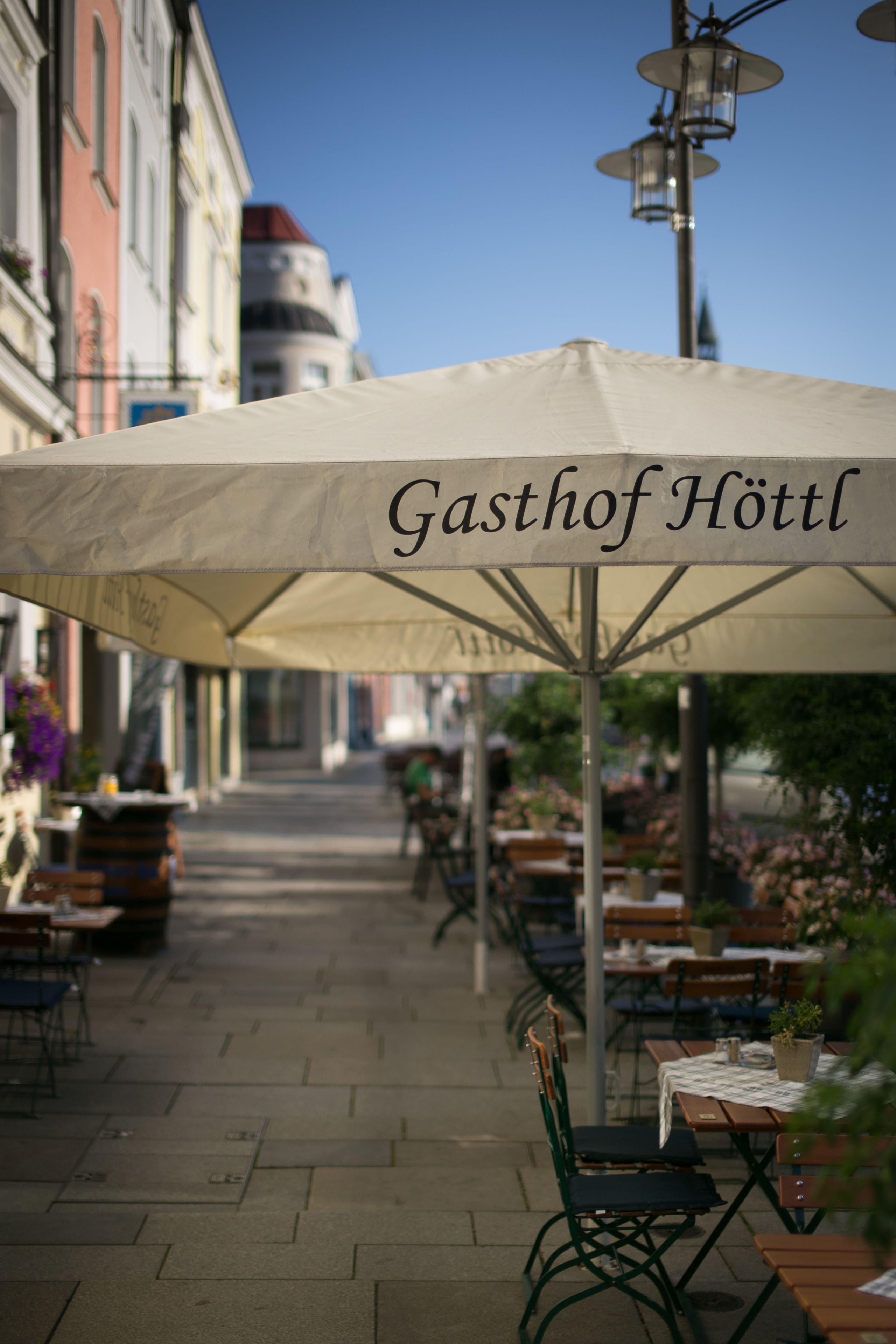 An outdoor café with tables and chairs under a large umbrella with the text "Gasthof Hötl".