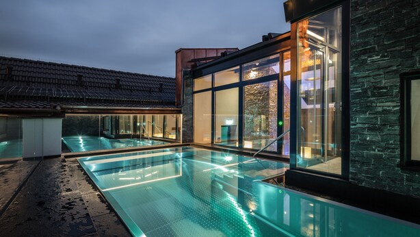 Una piscina exterior al lado de un edificio con grandes ventanales y paredes de piedra.