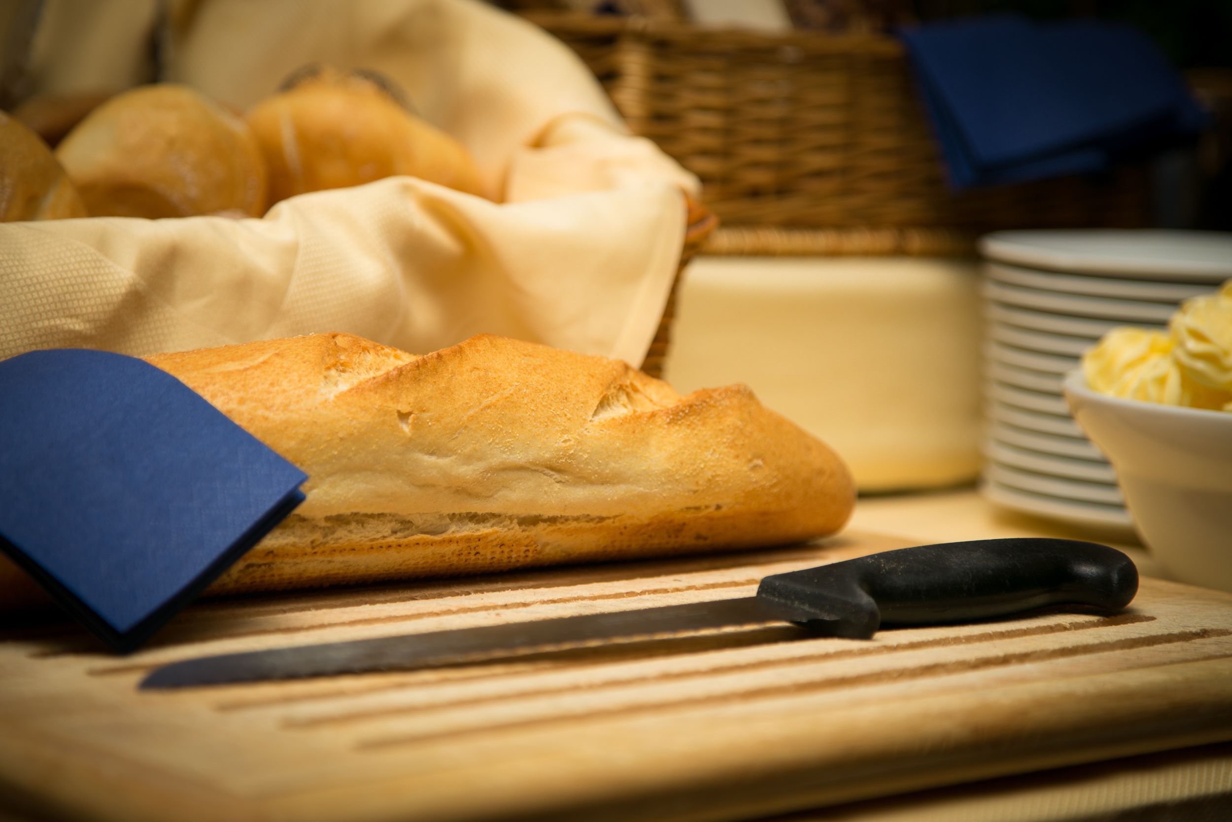 A loaf of bread on a cutting board with a knife, a basket of rolls, and a stack of plates.