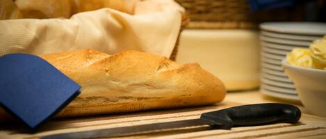 Une miche de pain sur une planche à découper avec un couteau, un panier de petits pains et une pile d’assiettes.